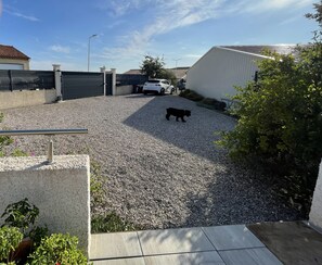 A gravel driveway with a dog walking, a white car parked, and a building with a sloped roof. - L'Olivier – A quiet, air-conditioned vacation rental in a traditional village near Pézenas (Neffiès)