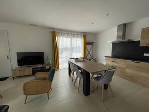 A modern living room with a flat-screen TV, a dining table with chairs, and a kitchen area with a stove and cabinets. - L'Olivier – A quiet, air-conditioned vacation rental in a traditional village near Pézenas (Neffiès)