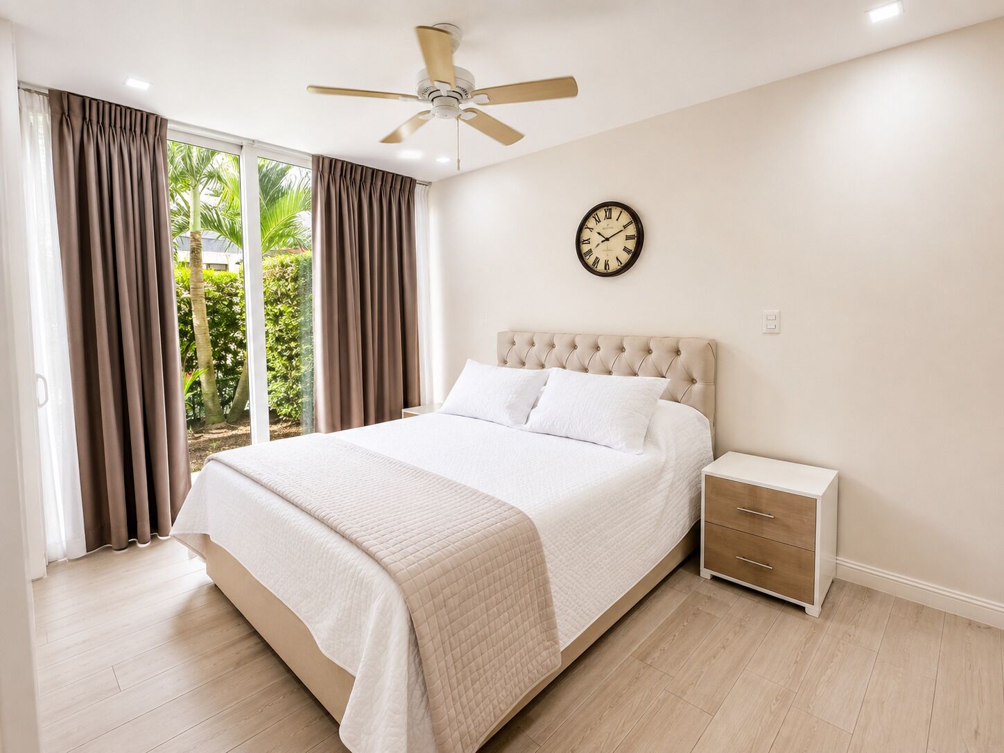Ein Schlafzimmer mit einem Bett, einem Nachttisch, einem Deckenventilator, einer Uhr und Blick auf Grünflächen.