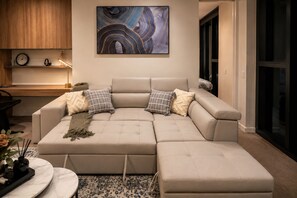 A modern living room with a sectional sofa, a large abstract painting, and a small wooden table with a clock and decorative items.