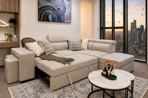 A modern living room with a sectional sofa, a round coffee table, and a city view through large windows. - 19F Sky-Rise 2BR 2BTH, Panoramic CBD Views with Resort-Style Amenities (Carlton)