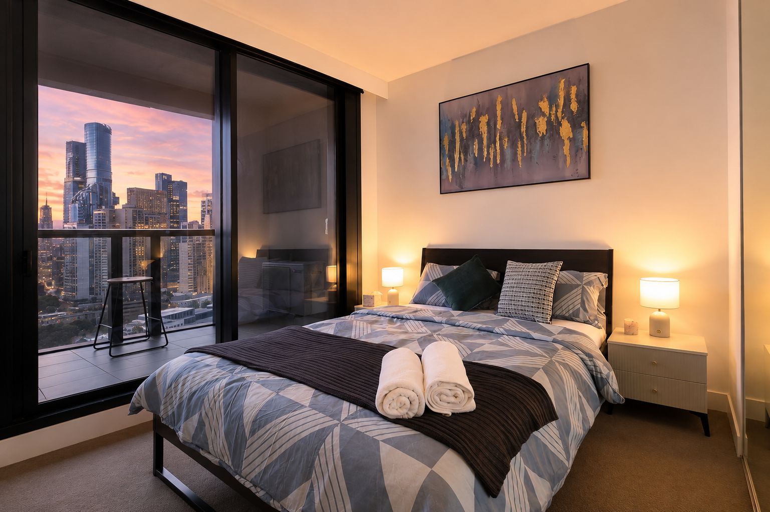 A hotel room with a large bed, two bedside tables, a city view, and a painting on the wall.
