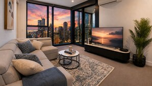 Un salon moderne doté d’un grand téléviseur, de sièges confortables, d’une table basse et offrant une vue sur les toits d’une ville au coucher du soleil.