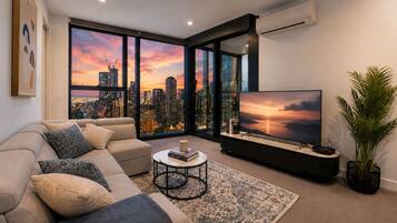 A modern living room with a large TV, comfortable seating, a coffee table, and a view of a city skyline at sunset.