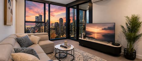 A modern living room with a large TV, comfortable seating, a coffee table, and a view of a city skyline at sunset.