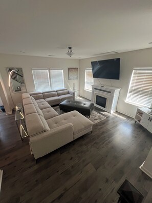 A modern living room with a sectional sofa, a flat-screen TV mounted above a fireplace, and a staircase with a railing. - Tidy 3BR High Point Retreat HPU-Furniture Market-Winston (High Point)