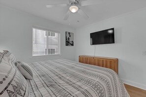 Un dormitorio con cama, ventilador de techo, televisor montado en la pared y ventana con persianas.