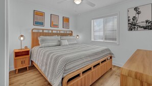 A bedroom with a bed, bedside tables, a ceiling fan, a window with blinds, and wall art. - Beach Getaway | Updated 2BR Condo, Pool & Beach Access (Carolina Beach)