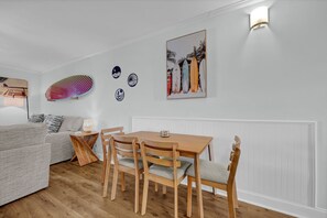 Ein gemütlicher Wohnbereich mit einer Couch, einem Esstisch mit Stühlen, einer Lampe und Wanddekorationen, darunter Surfbretter und ein gerahmtes Bild.