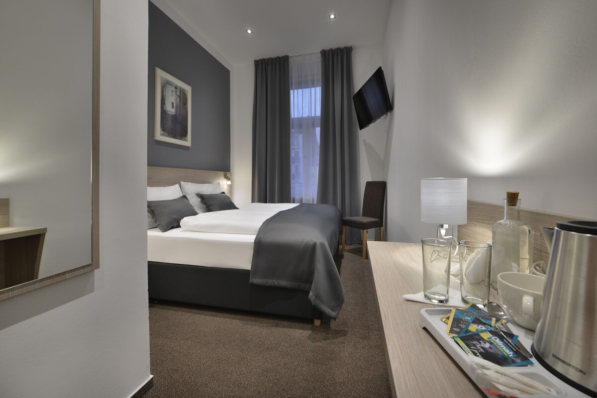 A hotel room with a bed, a desk with a lamp, a chair, a TV mounted on the wall, and a window with curtains.