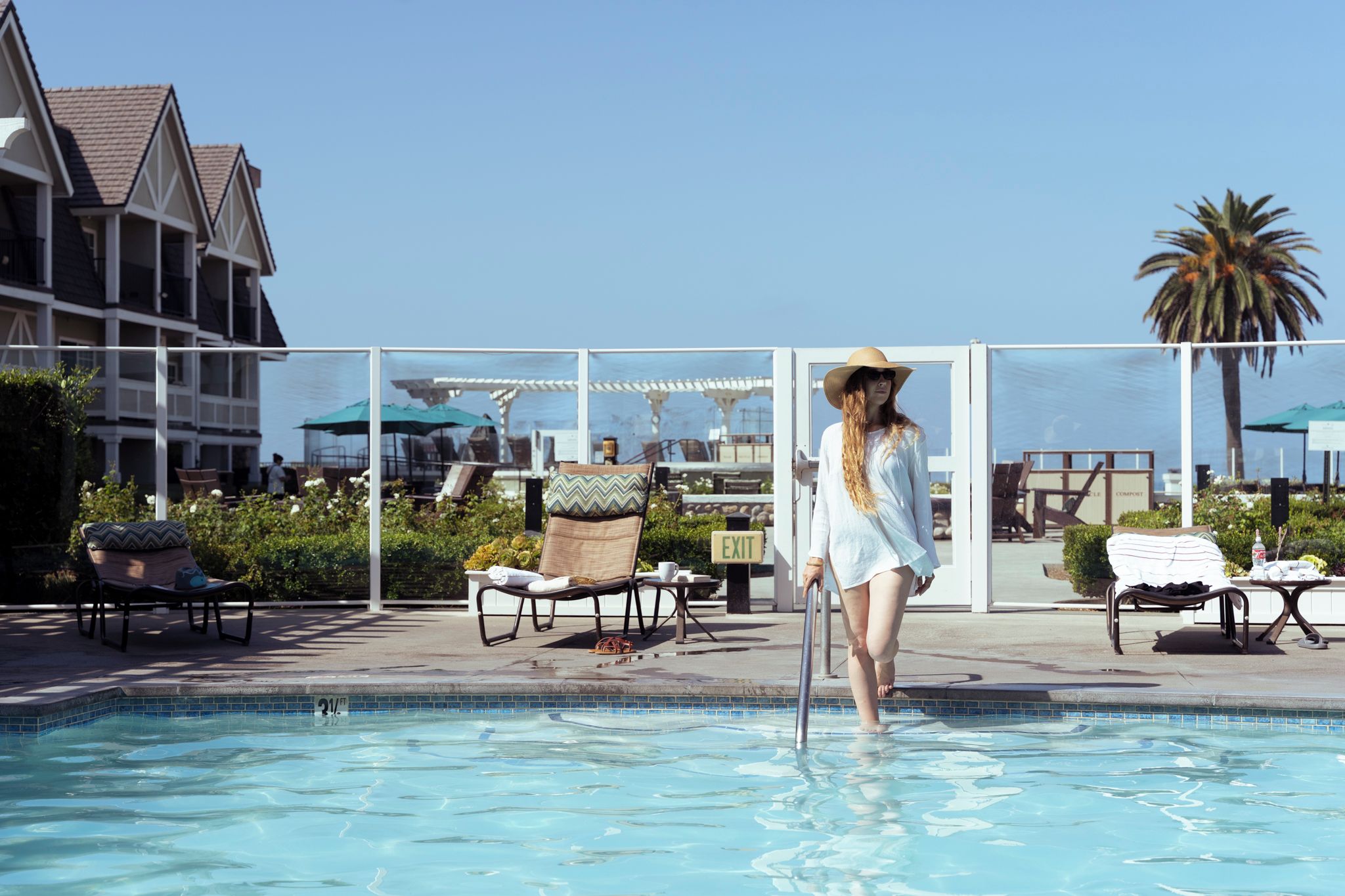 Zona de piscina con sillones de descanso y sombrillas, cielo despejado y un edificio al fondo.