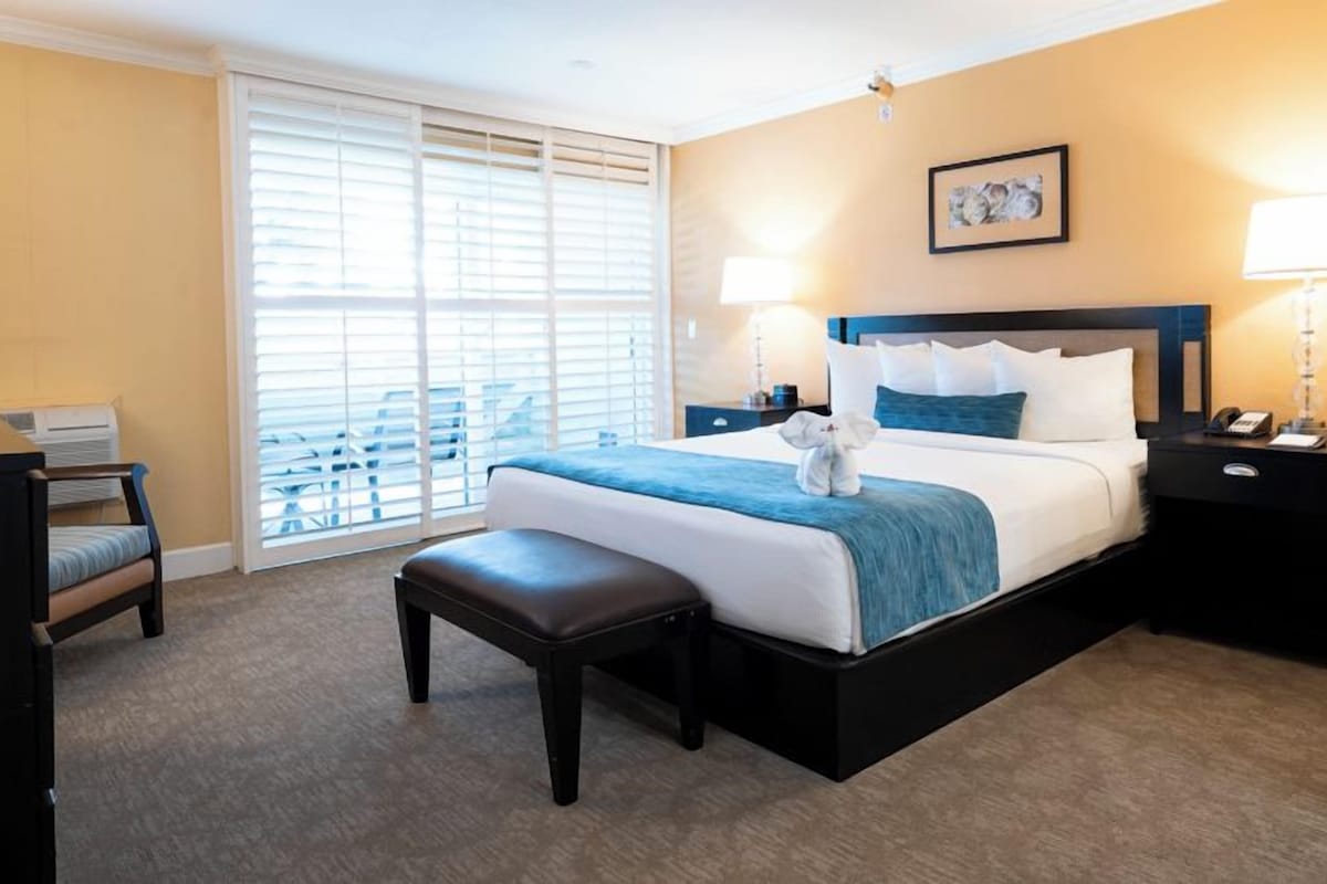 A hotel room with a bed, bedside tables, a bench, a desk, and a window with blinds.