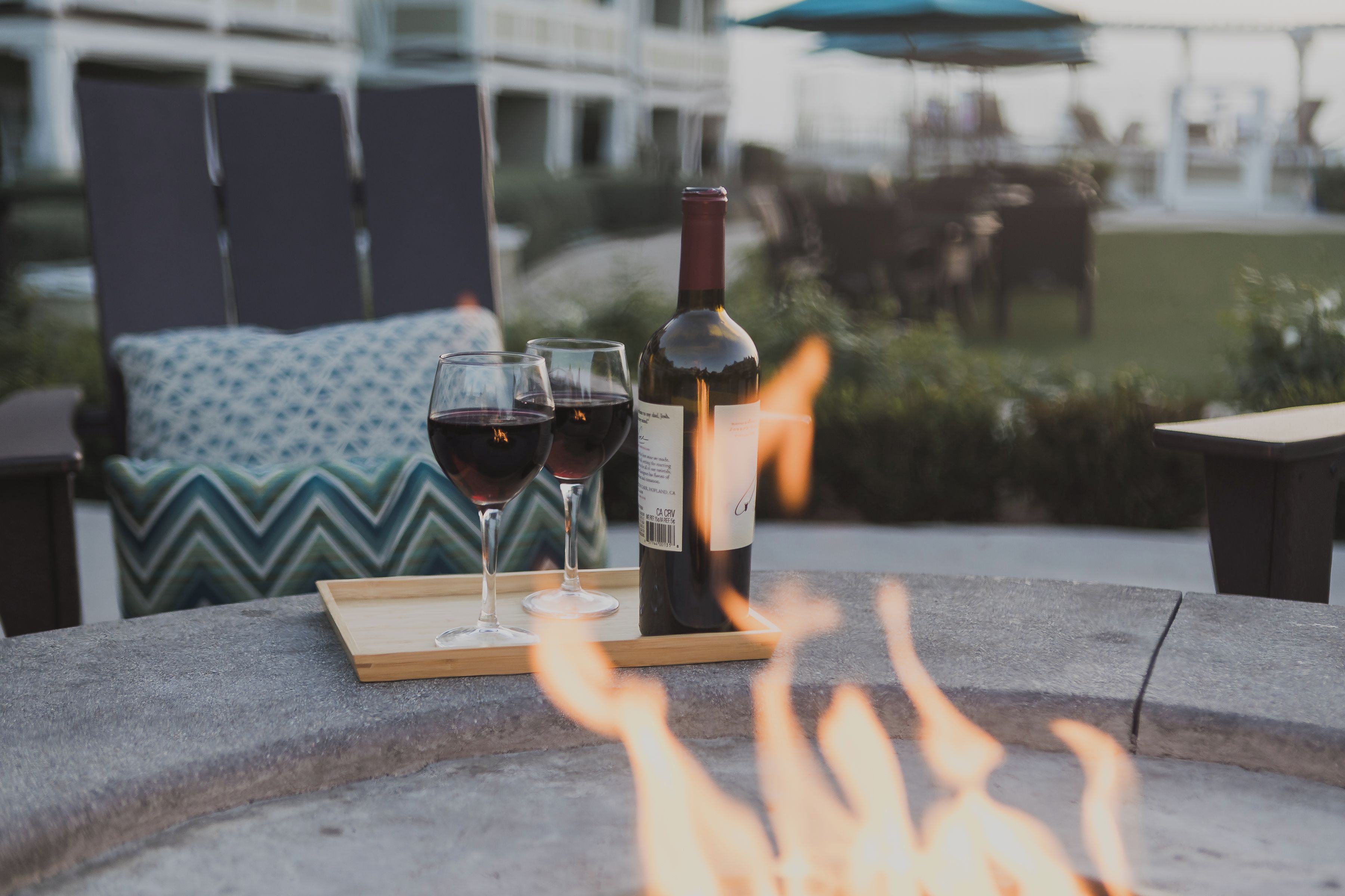 A fire pit with two glasses of red wine and a bottle on a wooden tray.