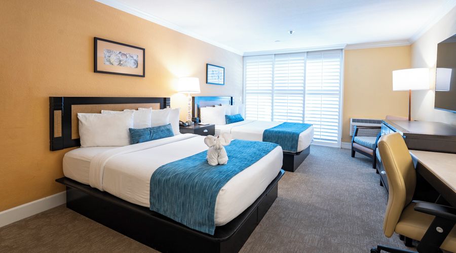 A hotel room with two beds, a desk, a chair, and a large window with blinds.