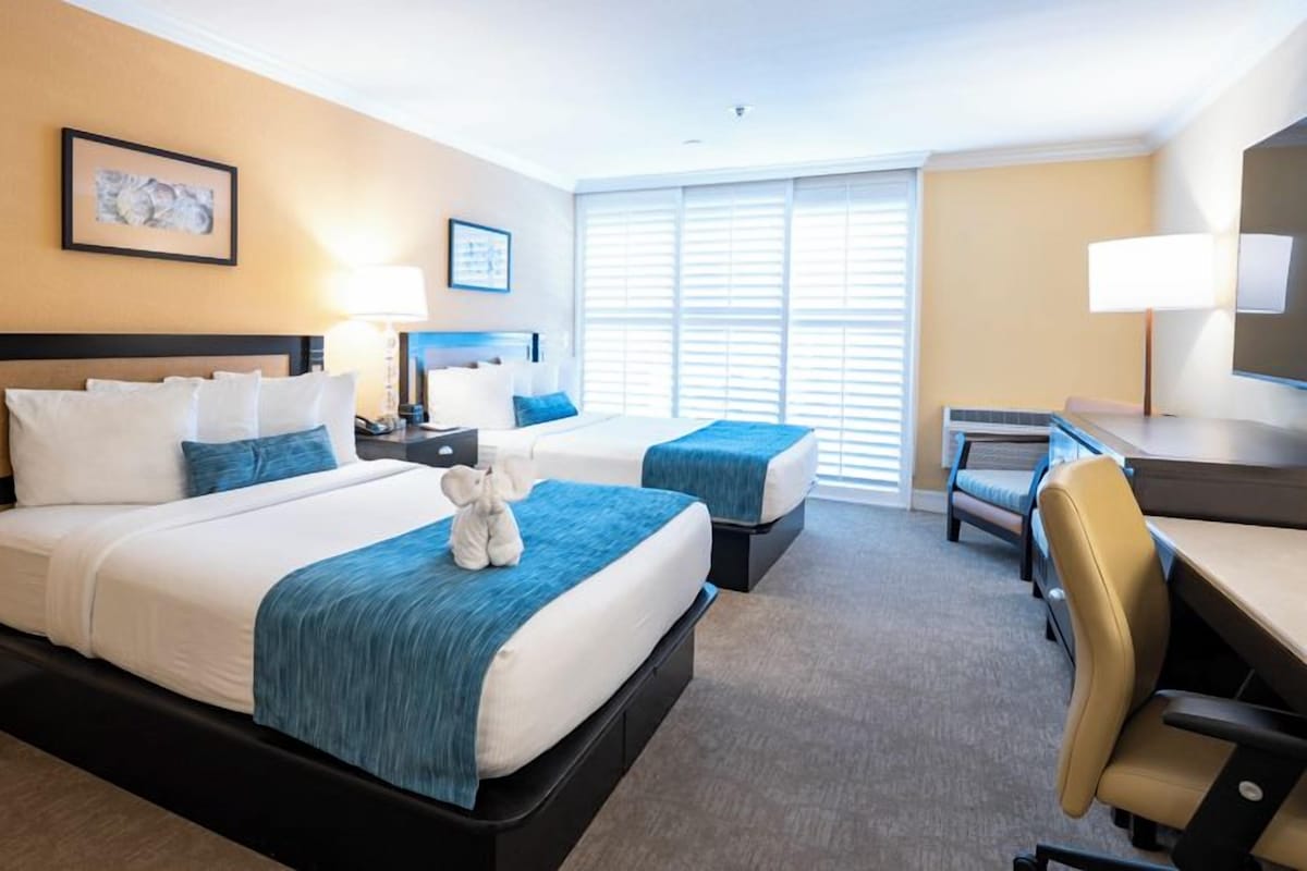 A hotel room with two beds, a desk, a chair, and a large window with blinds.