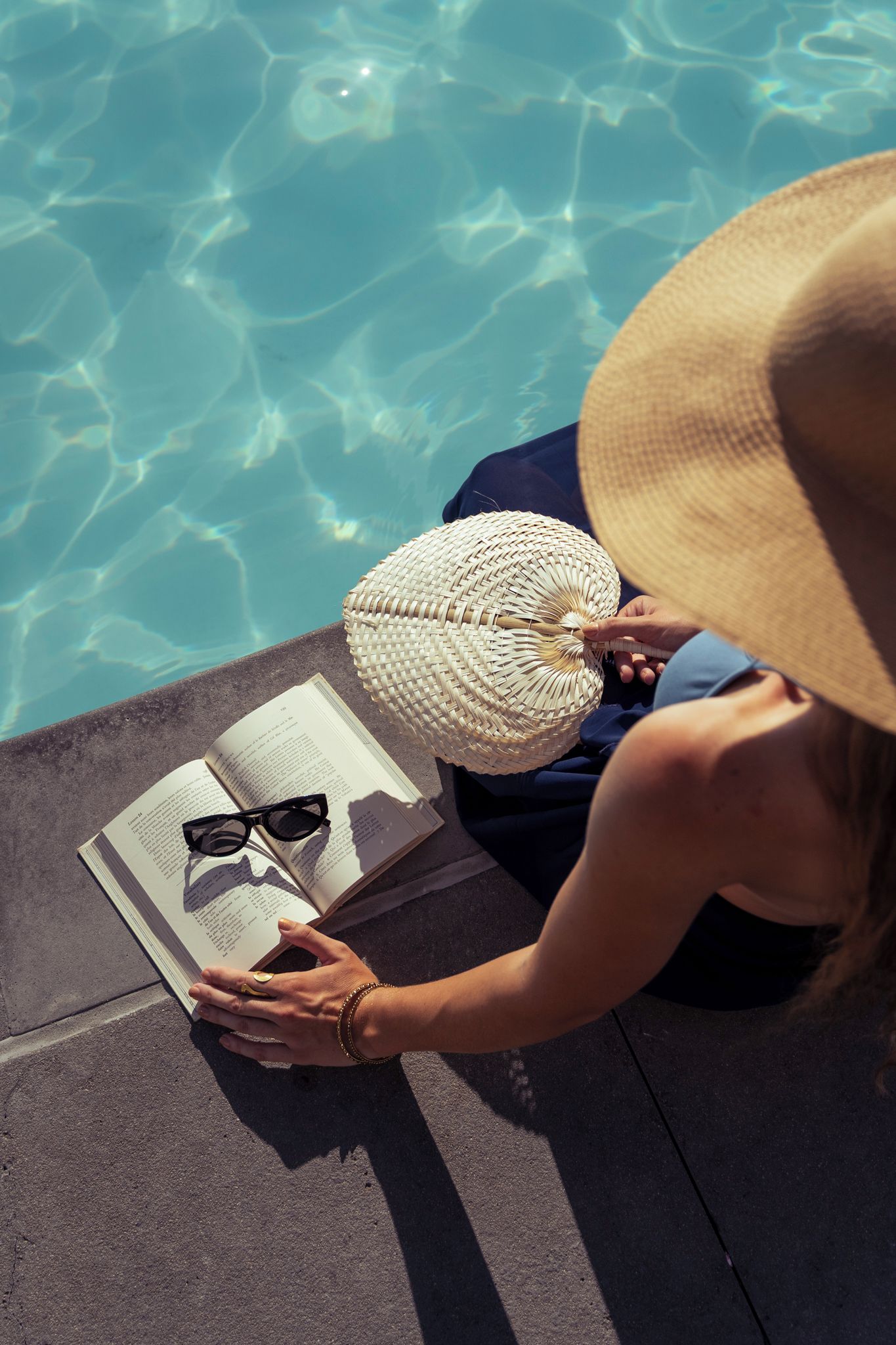 A person reading a book by a pool with a straw hat and sunglasses.