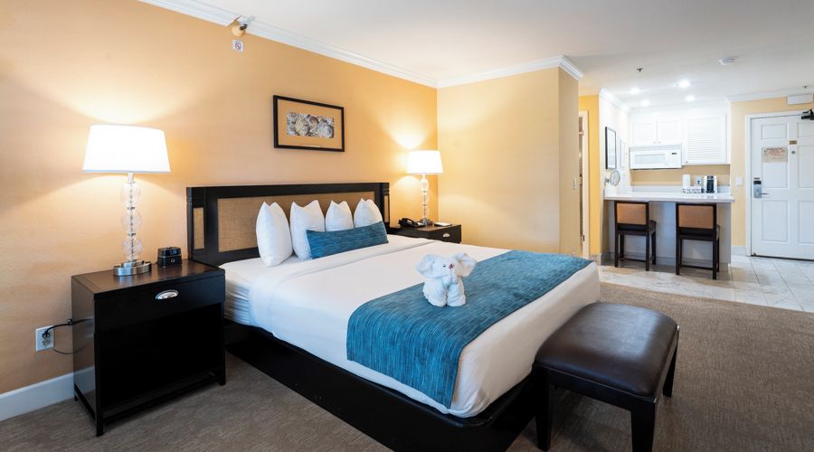 A hotel room with a bed, bedside tables, lamps, a small desk, and a kitchenette area.
