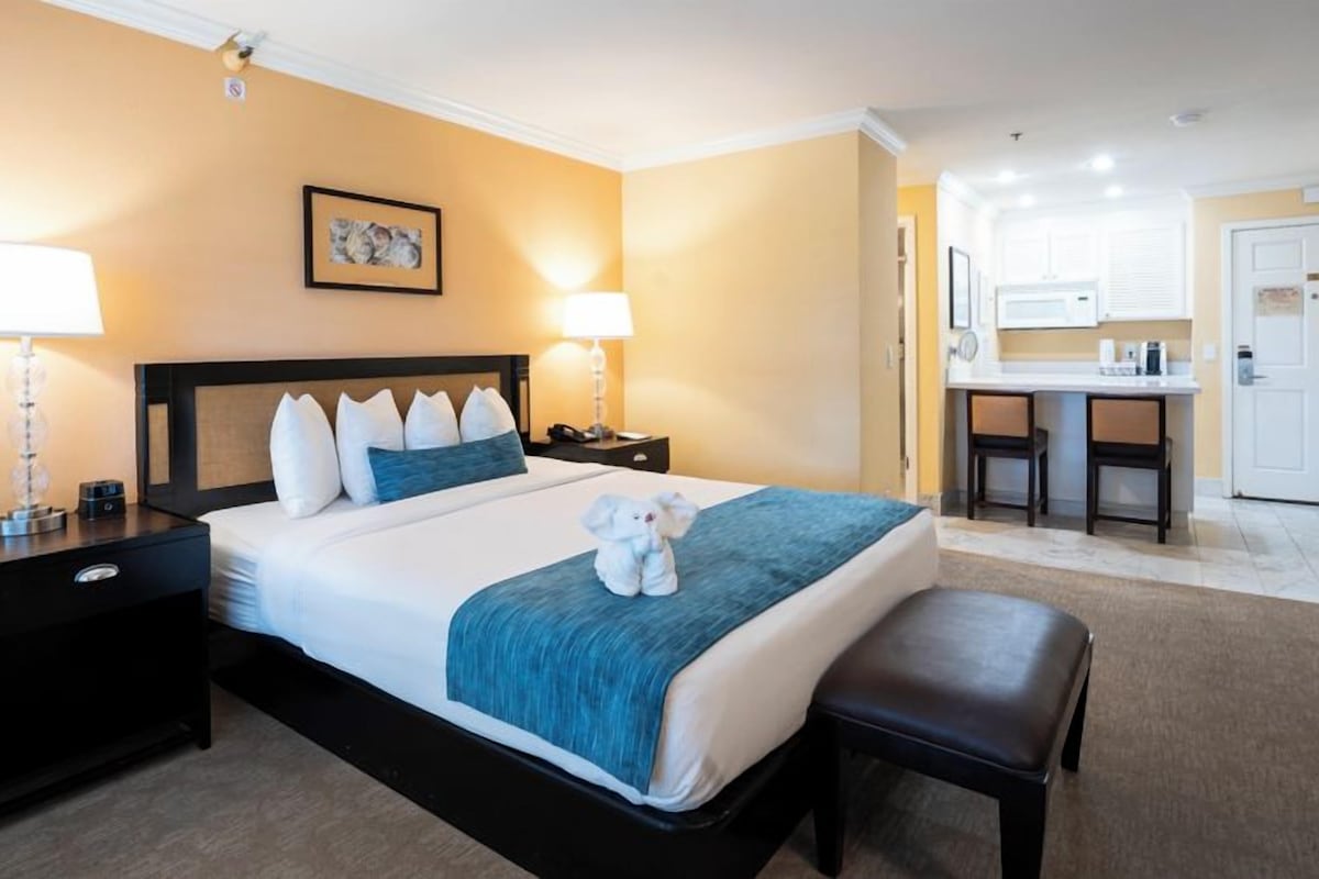 A hotel room with a bed, bedside tables, lamps, a small desk, and a kitchenette area.