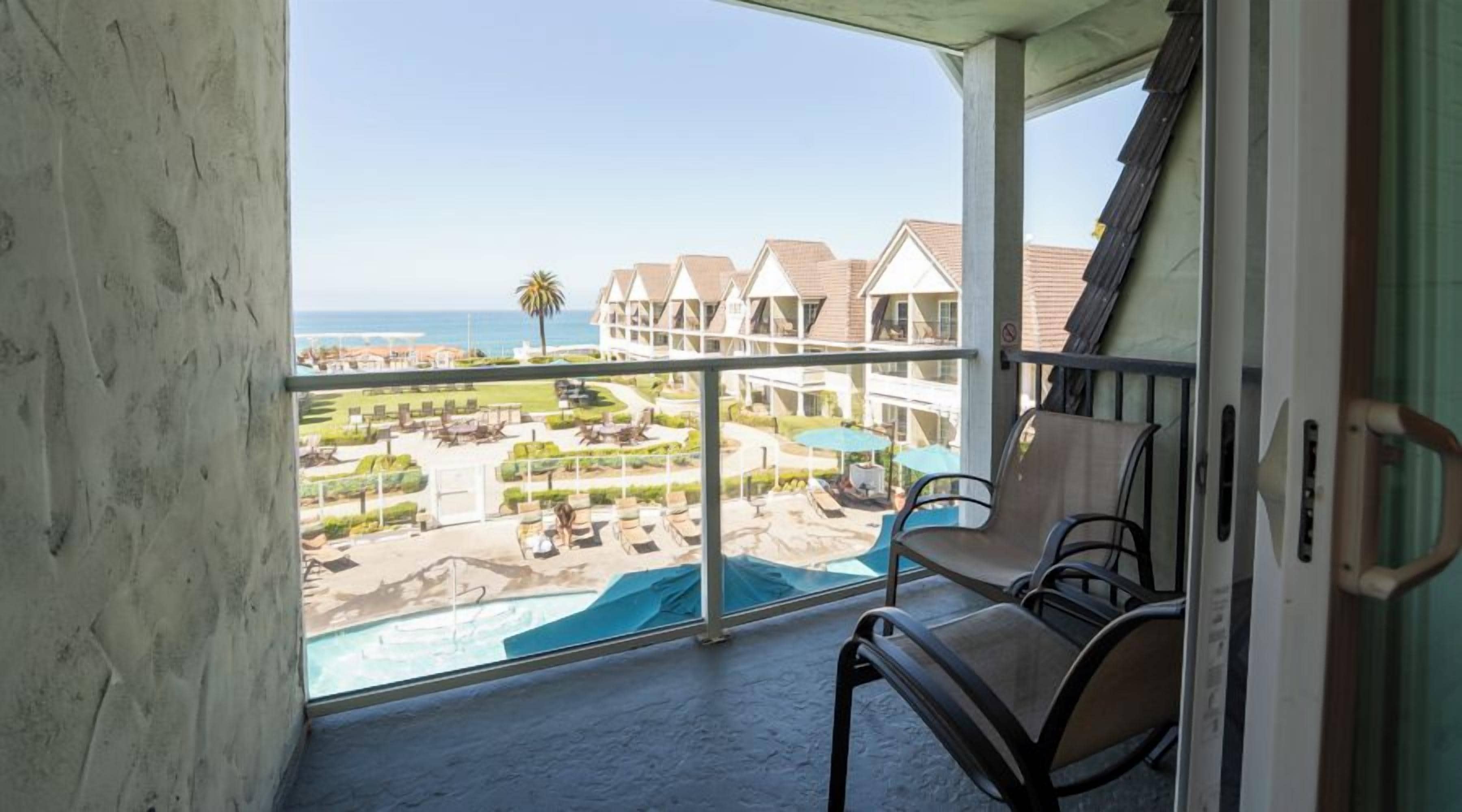 Un balcón con vistas a una piscina y zona de playa, con sillones de descanso y sombrillas.