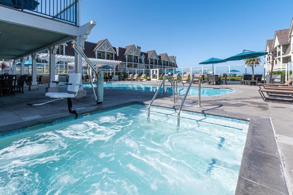 Ein Schwimmbecken mit Liegestühlen und Sonnenschirmen, umgeben von Gebäuden mit einer überdachten Terrasse.