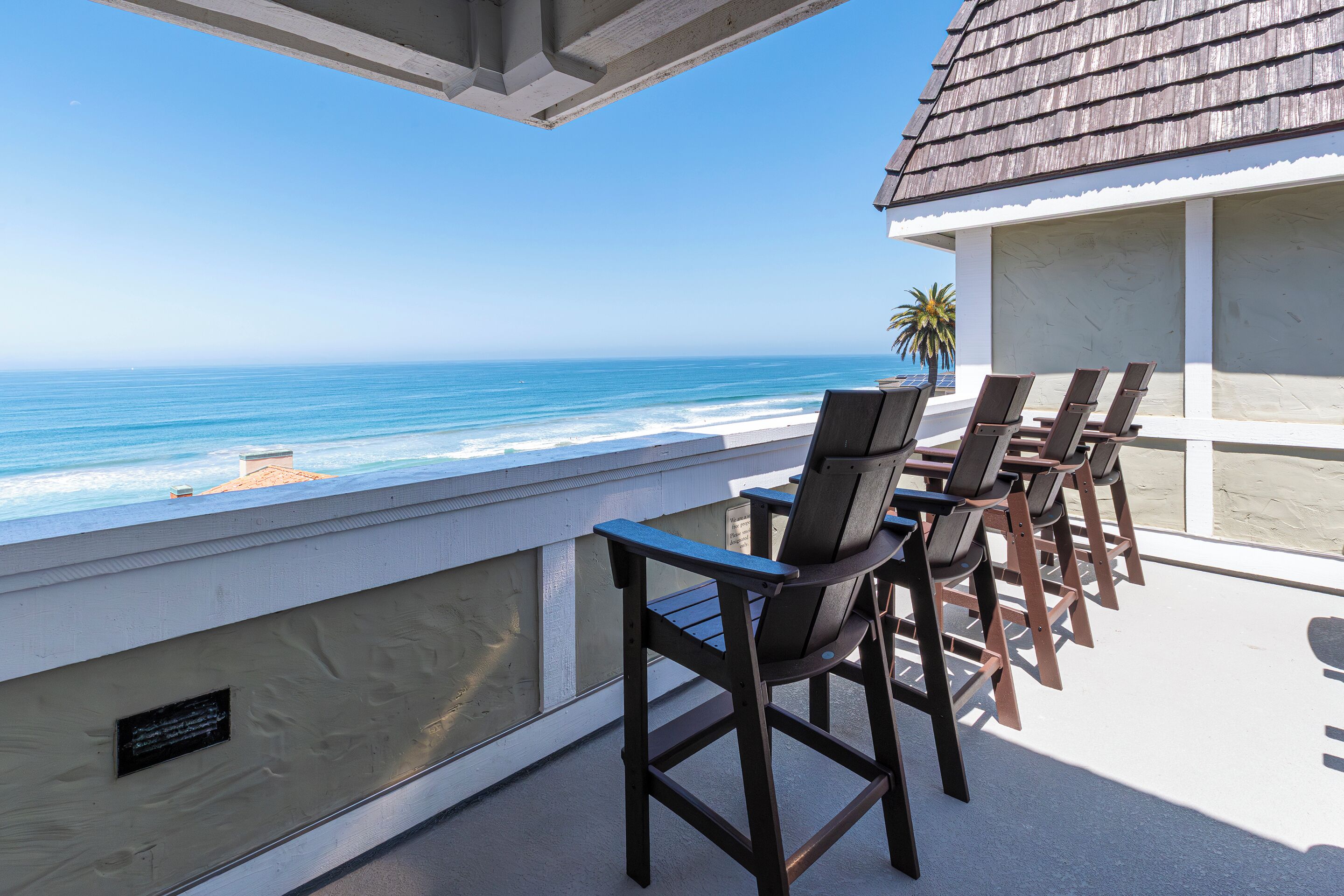 A balcony with a view of the ocean, wooden chairs, and a palm tree.
