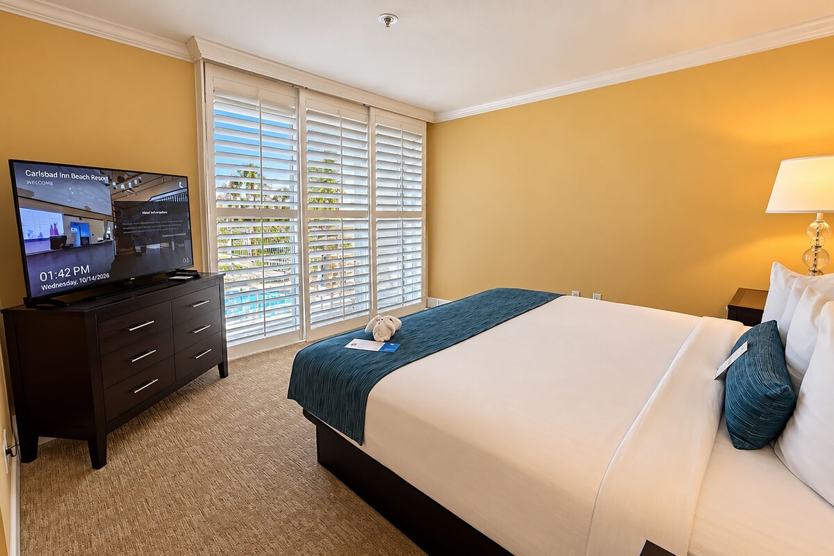 A hotel room with a bed, a TV on a dresser, and a window with blinds.