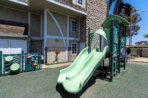 A playground with a green slide and climbing structures, located in front of a building with a stone facade and a gated entrance. - Carlsbad Inn Beach Resort Hotel (Carlsbad)