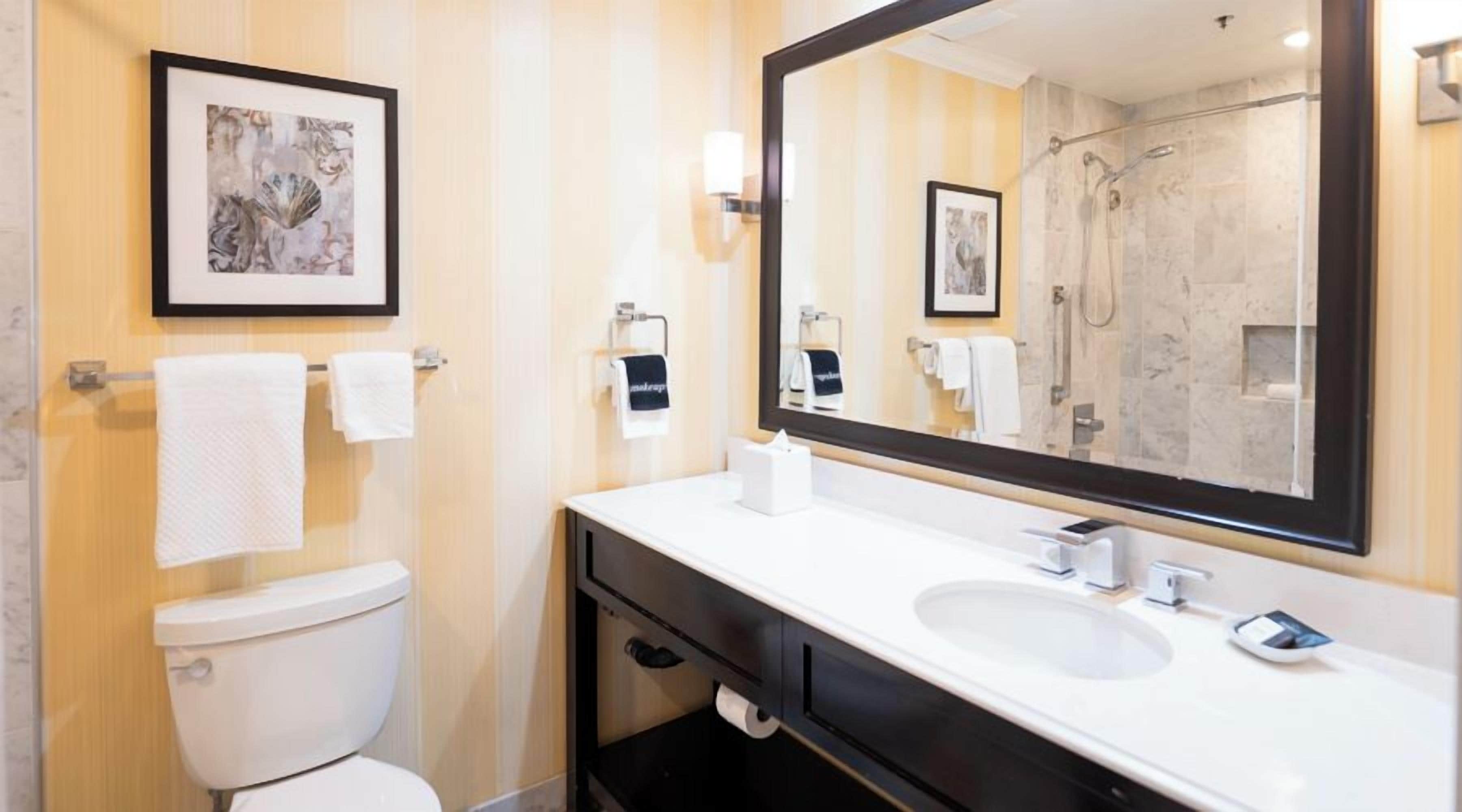 A modern bathroom with a toilet, sink, and shower. There are framed pictures on the wall and a large mirror above the sink.