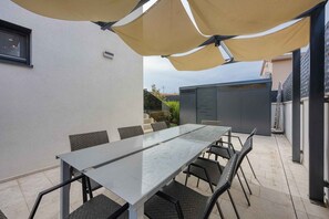Eine Terrasse im Freien mit einem rechteckigen Tisch und Stühlen, umgeben von einem beigefarbenen Sonnensegel.