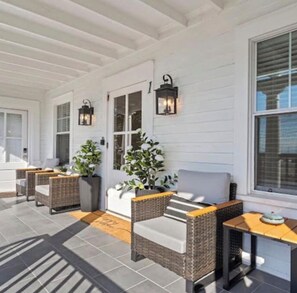 A covered outdoor seating area with wicker furniture, a sofa, and a table, featuring a white exterior wall and a window with blinds.