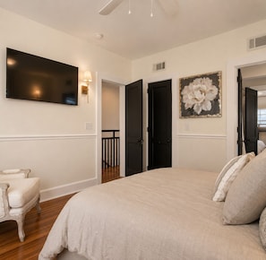 A bedroom with a large bed, a flat-screen TV mounted on the wall, a door leading to another room, and a painting on the wall.