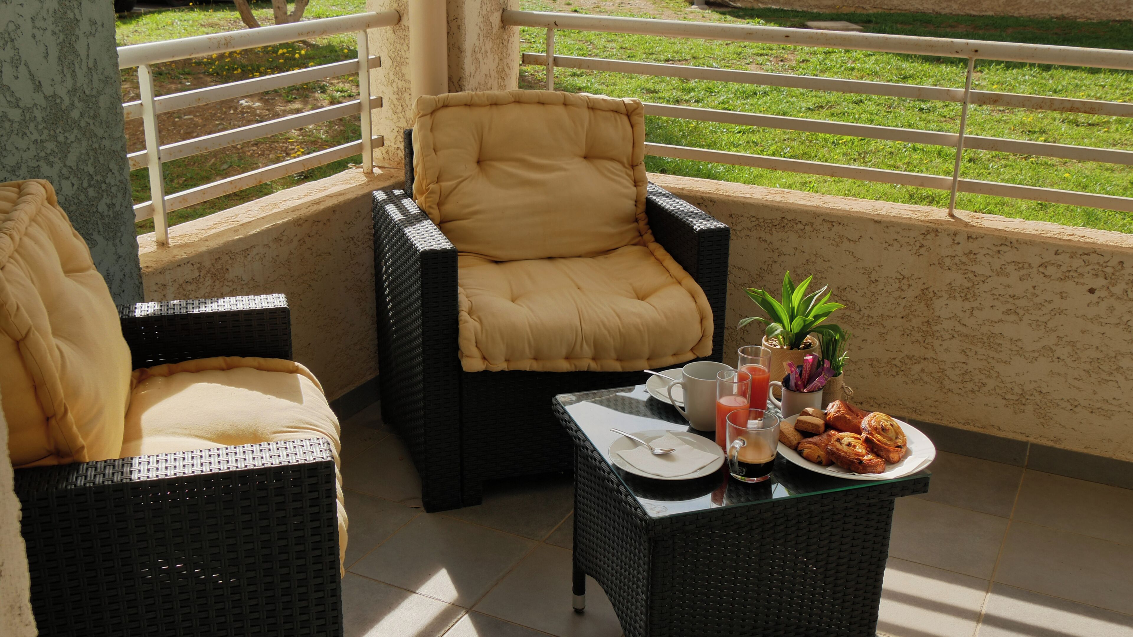A balcony with wicker furniture, a glass table, and a view of a grassy area.