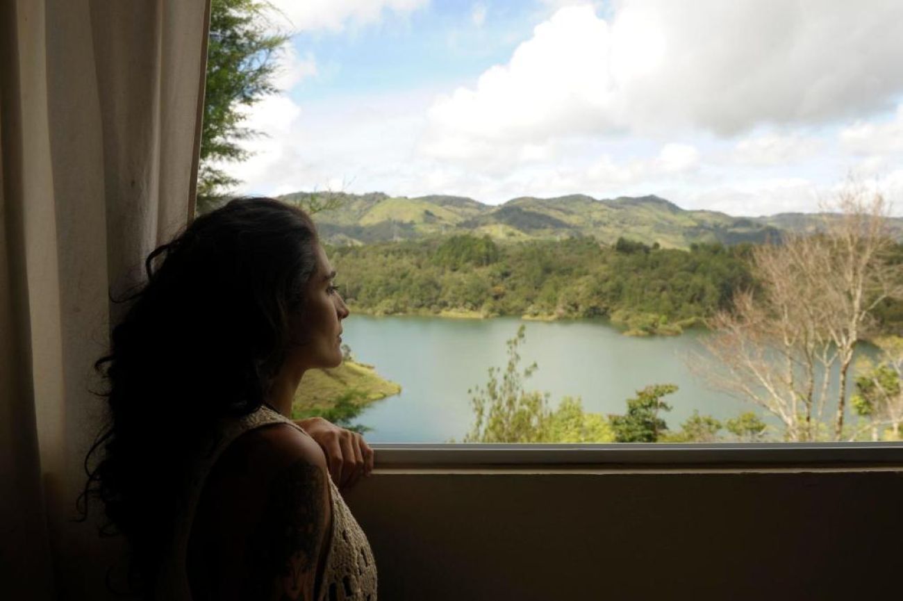 Alguém observa a paisagem pela janela, com vista para um lago e montanhas.