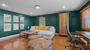 Ein Wohnzimmer mit einem weißen Sofa, einem Holztisch und einem braunen Ledersessel. An der Wand hängen Bilderrahmen und es gibt ein Fenster mit Jalousien.