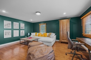 Ein Wohnzimmer mit einem weißen Sofa, einem Holztisch und einem braunen Ledersessel. An der Wand hängen Bilderrahmen und es gibt ein Fenster mit Jalousien.