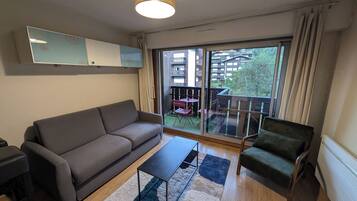 Un salon moderne avec un canapé, un fauteuil et une table basse. Une porte-fenêtre coulissante donne accès à un balcon extérieur équipé d’un ensemble de mobilier de jardin.