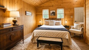 Quarto aconchegante com teto de madeira, cama com colcha acolchoada, luminárias de cabeceira, uma cadeira e cômoda.
