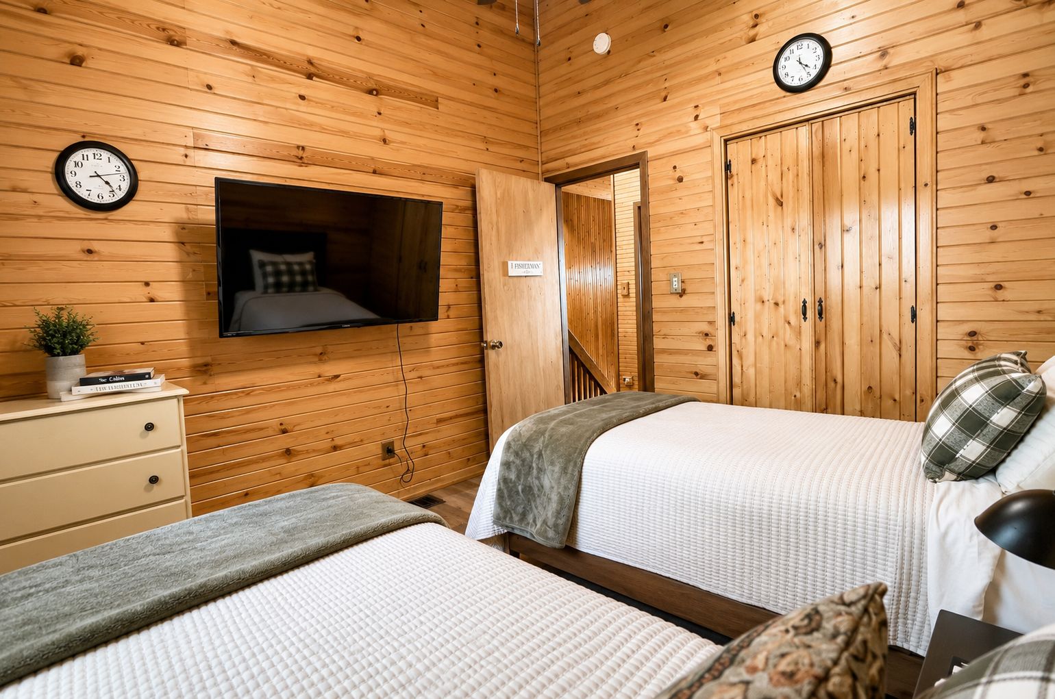 Ein Zimmer mit Holzwänden, zwei Betten mit weißer Bettwäsche, einem an der Wand montierten Fernseher, einem Nachttisch mit einer Pflanze und einer Wanduhr.