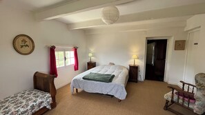 Ein Schlafzimmer mit einem Bett, zwei Nachttischen, einem Sessel, einem kleinen Tisch, einem Fenster mit Vorhängen und einer Tür.