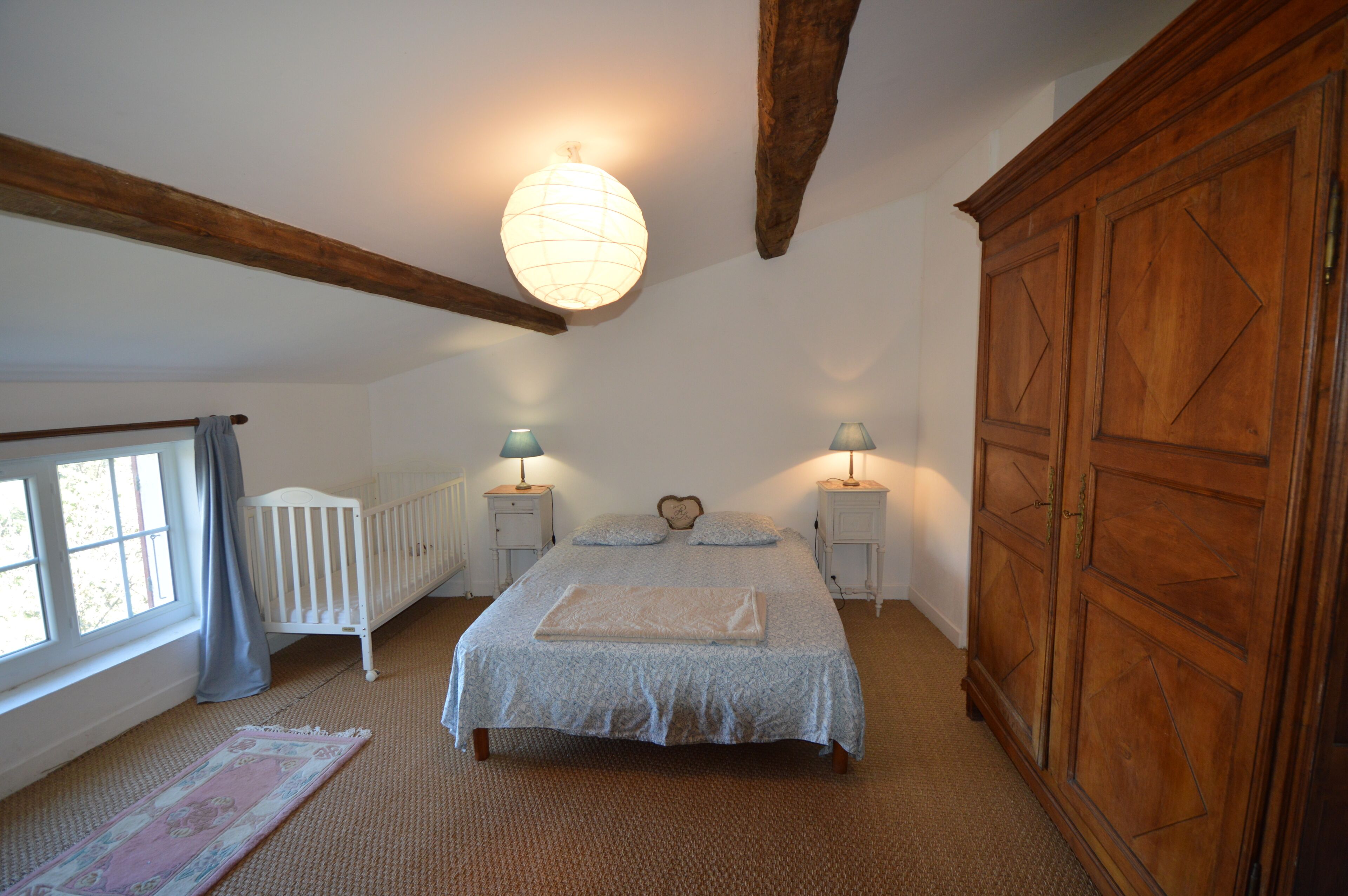 A bedroom with a large bed, two bedside tables with lamps, a wooden wardrobe, a radiator, and a window with curtains.