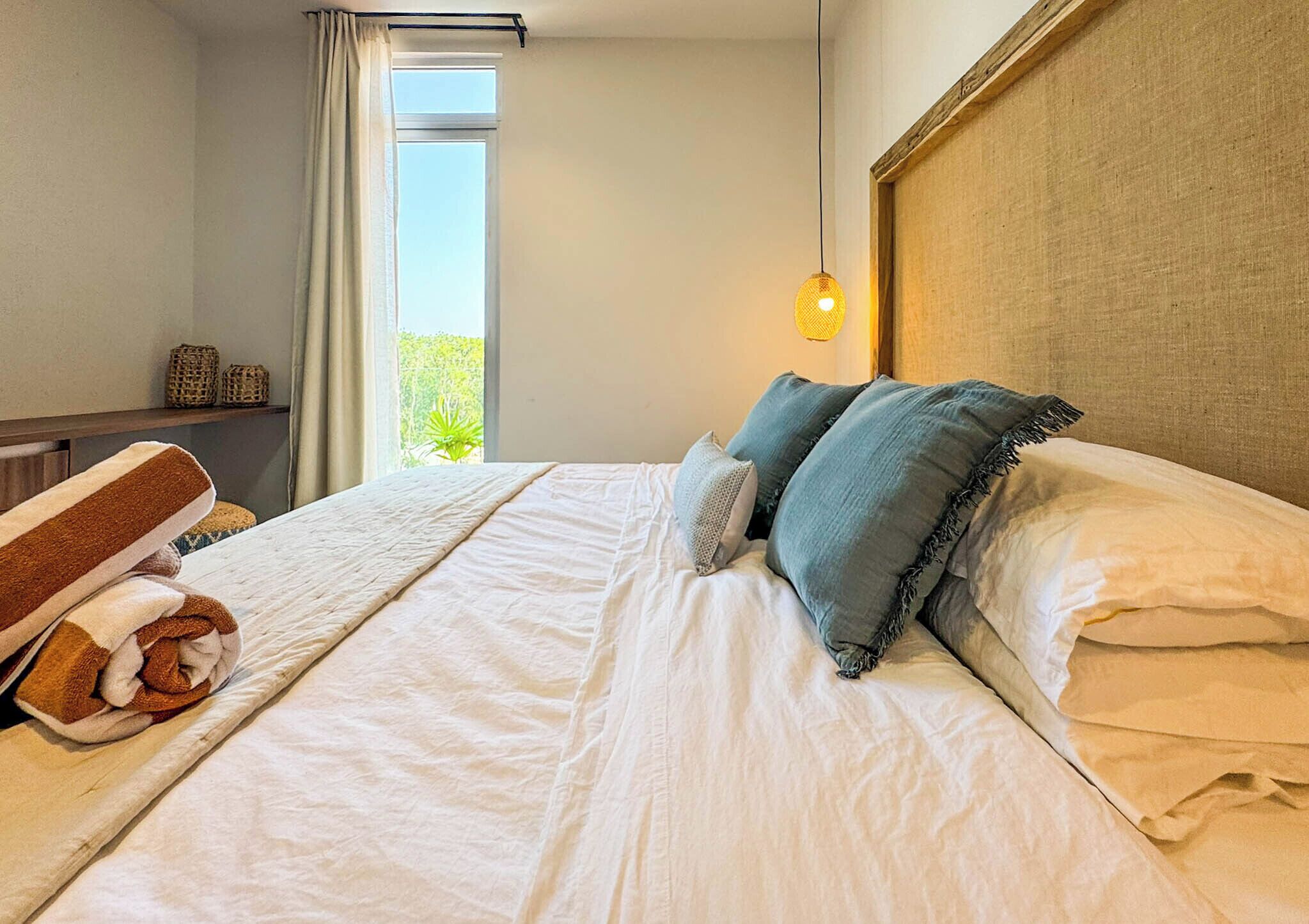 Een hotelkamer met een bed, kussens, een nachtkastje, een lamp en een raam met gordijnen.