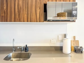 A kitchen with a stainless steel sink, a microwave, wooden cabinets, a soap dispenser, a paper towel holder, and a stone mortar. - 3BR Tulum Condo with Garden Views (Tulum)