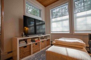 A room with a flat-screen TV on a white stand, a bookshelf with books and baskets, a window with blinds, and a bed with a white cover. - Walk to the Beach ~ 2-Bed Kinney Shores Retreat! (Saco)