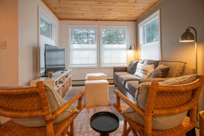 A cozy living room with a television, wicker chairs, a sofa, and a lamp. - Walk to the Beach ~ 2-Bed Kinney Shores Retreat! (Saco)