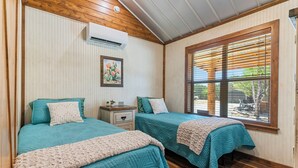 A bedroom with two beds, a nightstand, a wall-mounted air conditioner, and a window with blinds.
