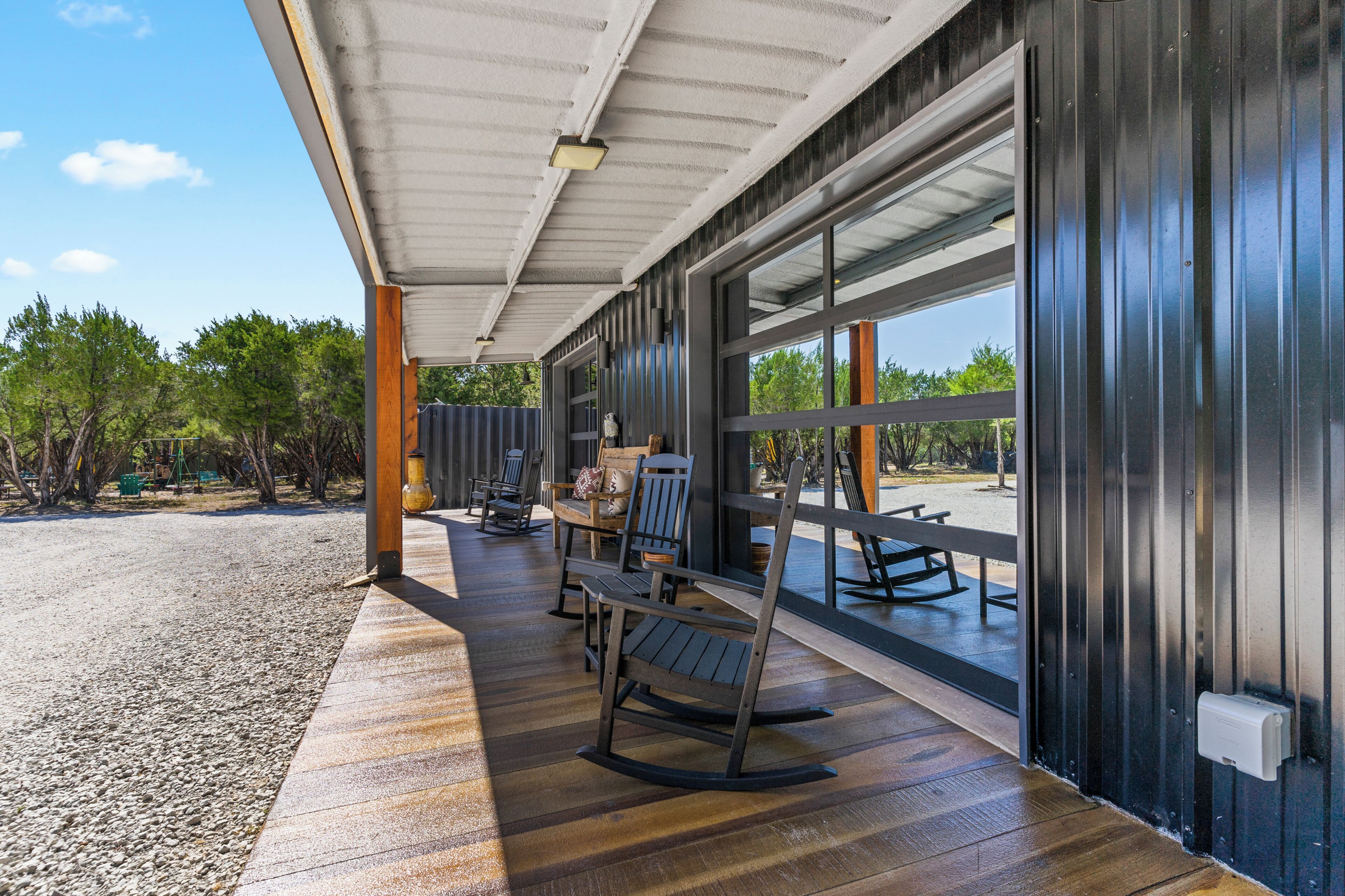 Un patio exterior con piso de madera, un área de descanso con sillas y una mecedora, y vistas a través de grandes ventanales.
