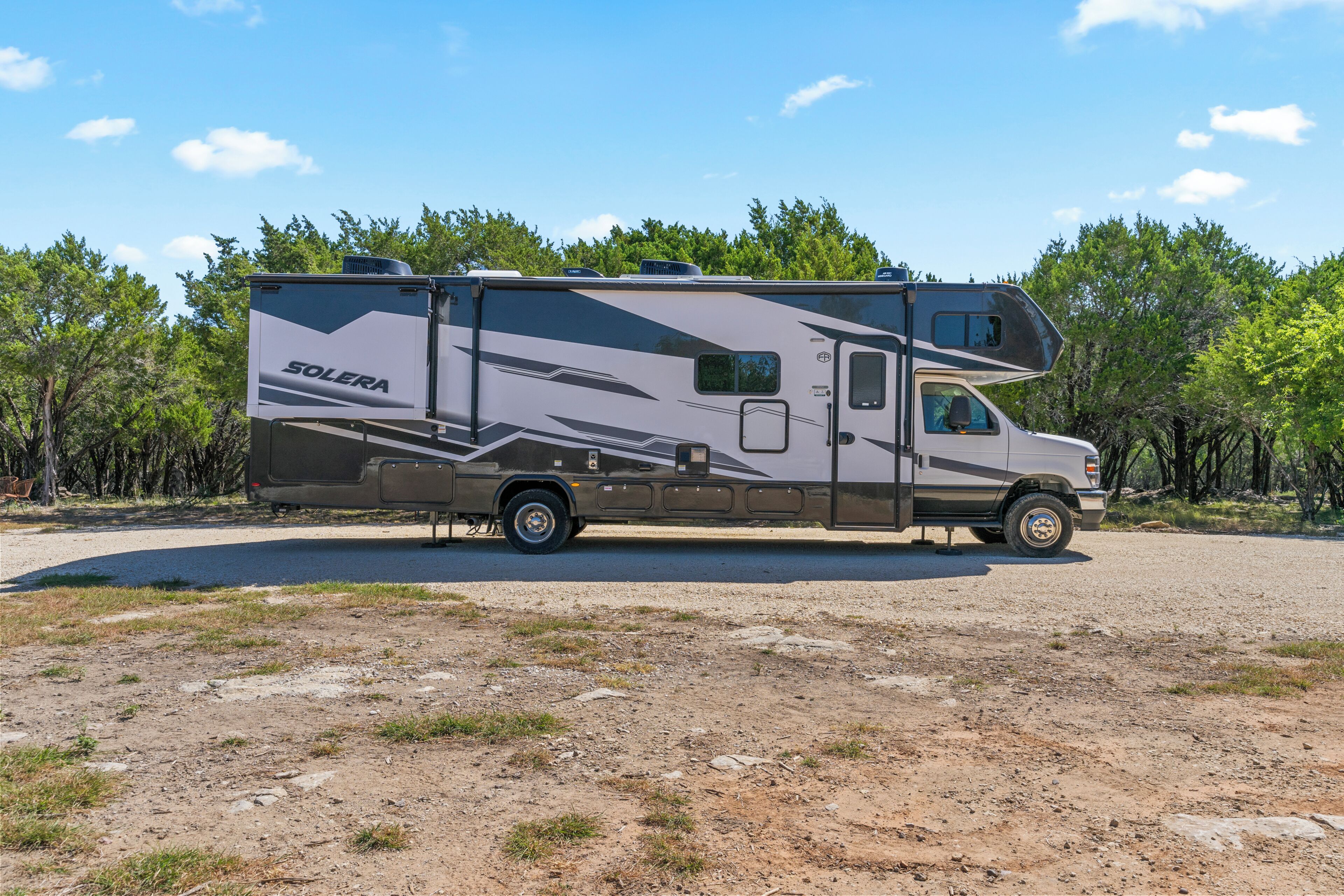Un grand camping-car doté d’une partie aménagée en espace de vie, garé sur un terrain recouvert de gravier, avec des arbres en arrière-plan.