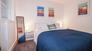 A bedroom with a bed, a nightstand, a mirror, and three framed posters on the wall. - Higher Tower Road (Newquay)