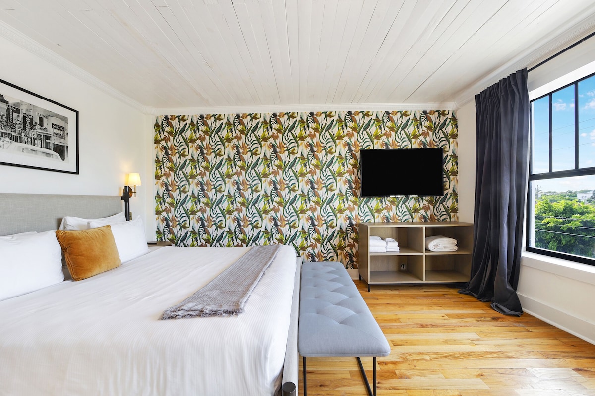 A hotel room with a bed, a bench, a TV, a shelf with towels, and a window with a view.