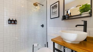 A modern bathroom with a white tiled shower, a wooden vanity with a white sink, and a framed picture on the wall.